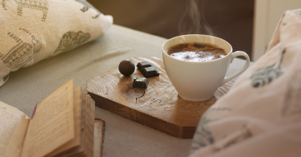 Hot coffee in a white mug with a nice smoke on the wooden coaster, dark chocolate pieces and an open book in bed